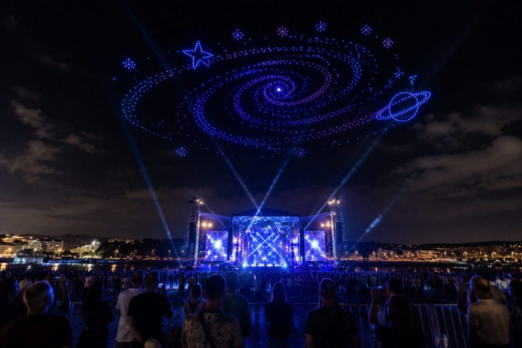 Show de drones lumineux au-dessus d’un concert en plein air avec une animation galaxie dans le ciel et une scène éclairée de nuit.