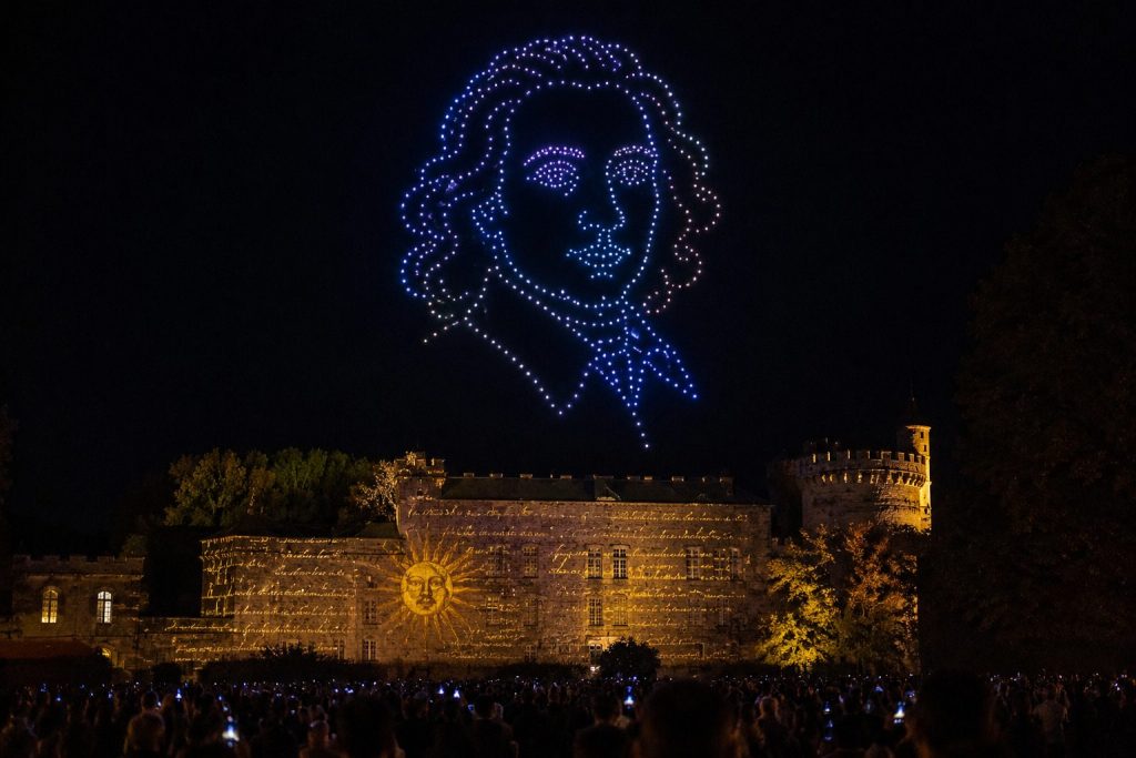 Spectacle de drones lumineux représentant un personnage historique au-dessus d’un château lors d’une célébration culturelle.